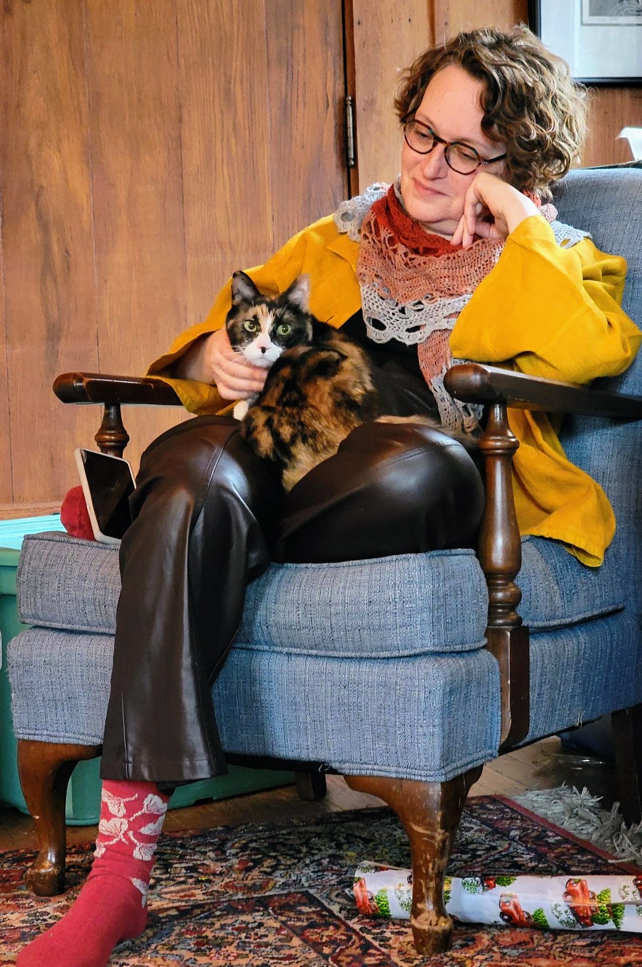 A white woman with curly short hair is wearing a mustard yellow linen shirt and a crocheted shawl in red's and greys. A calico cat sits on her lap and is wondering why someone is disturbing naptime with photos.