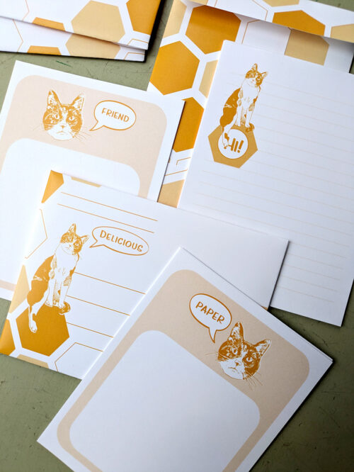 An array of stationary with orange details. A calico cat is on each. Bubbles say "friend" "Delicious" "Hi" and "Paper"
